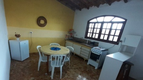 Cozinha com fogão, mesa, cadeiras, armário com pratos, copos, talheres, panelas e utensilios de coz 